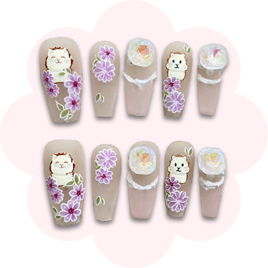 kawaii cat nail art press on nails on a pastel pink background with purple flower accents featuring  a rhinestone and pearls accents.